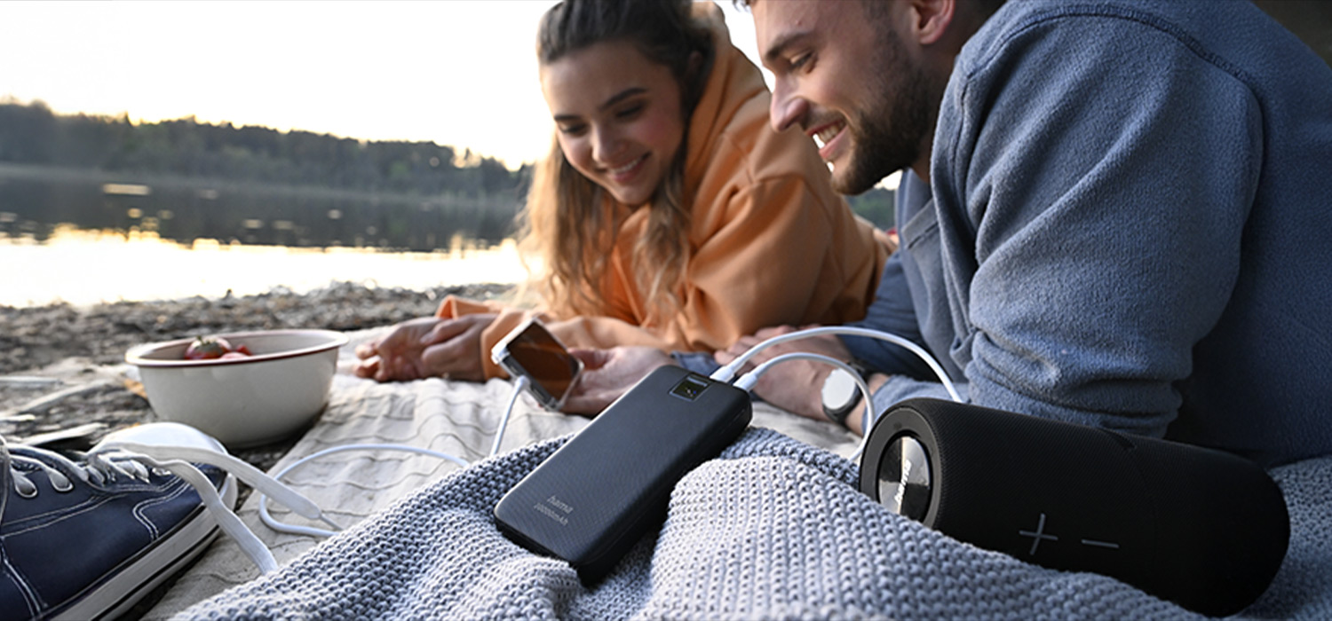 Ein Pärchen liegt am Badesee, im Vordergrund wird eine Boombox mit Energie von einer Powerbank gespeist.