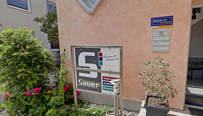 Gebäude mit Schild "Sauer", Pflanzen, Briefkasten, "Meisterhaft"-Schild und Eingangstür. Ein kleiner Baum steht in einem Topf.