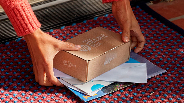 Eine Person in rotem Pullover nimmt eine braune Schachtel und Briefe vom roten Teppich vor einer Tür auf.
