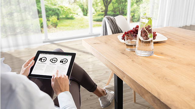 Eine Person bedient ein Tablet mit Hausautomations-Symbolen. Auf dem Tisch stehen Trauben und ein mit Wasser gefülltes Glas.