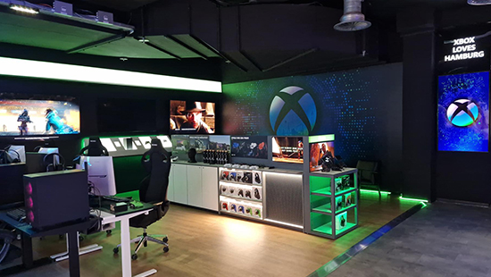 Ein Verkaufsraum mit Spielekonsolen, Bildschirmen und Zubehör, beleuchtet mit grünen Akzenten und dem Xbox-Logo.