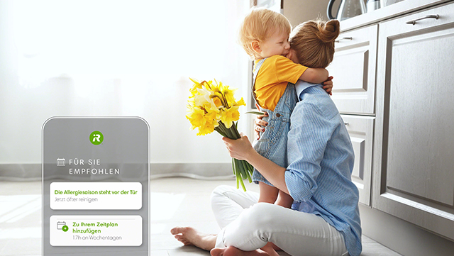 Frau umarmt Kind, hält gelbe Blumen. App-Interface zeigt 'Für Sie empfohlen'. Küche mit weißen Schränken im Hintergrund.