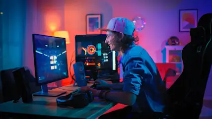 Ein Mann mit Kappe sitzt an einem Tisch mit Computermonitor und spielt vor einem beleuchteten Hintergrund.