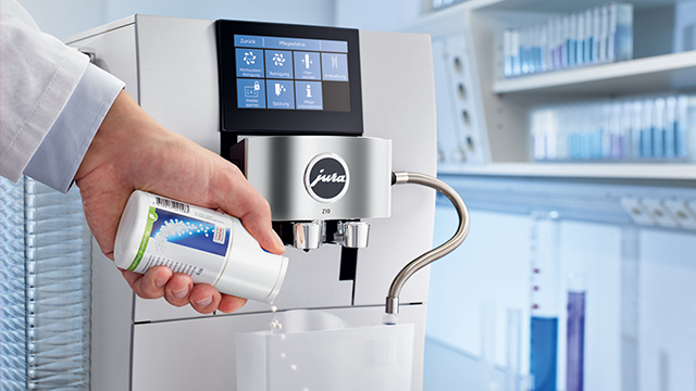 Eine Hand hält eine Filterpatrone über einen Jura-Kaffeevollautomaten. Im Hintergrund sind Regale mit Röhrchen sichtbar.