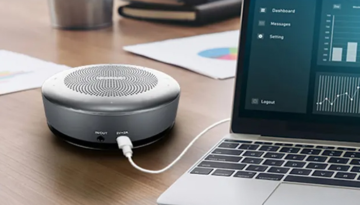 Ein grauer, runder Lautsprecher ist per Kabel mit einem geöffneten Laptop auf einem Holztisch verbunden. Diagramme sind auf dem Laptop-Bildschirm sichtbar.