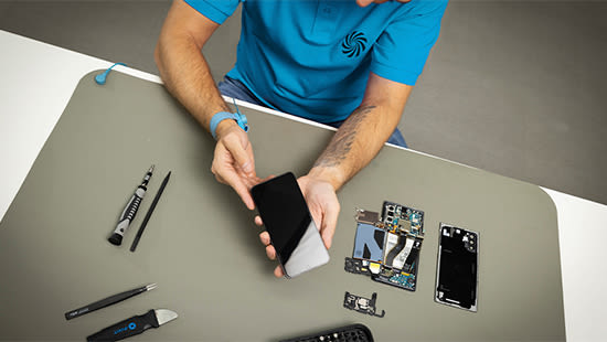Ein Mann im blauen T-Shirt repariert ein Smartphone. Das passende Werkzeug liegt neben ihm.
