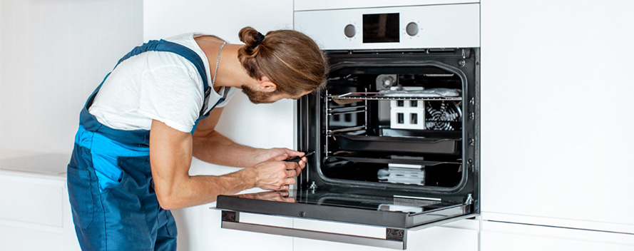Ein Mann in blauer Arbeitskleidung repariert einen weißen Backofen mit Gitter und Ventilator in einer weißen Küche.