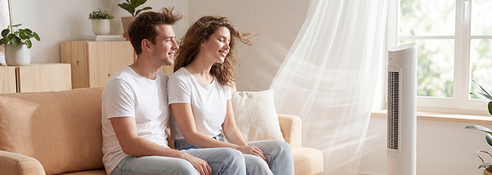 Junges Paar in weißen T-Shirts sitzt zusammen auf hellem Sofa, sichtbare Luftbewegung durch Ventilator. Weißer Ventilator im Vordergrund. Helles Wohnzimmer mit Holzmöbeln, Pflanzen und natürlichem Licht von großem Fenster.