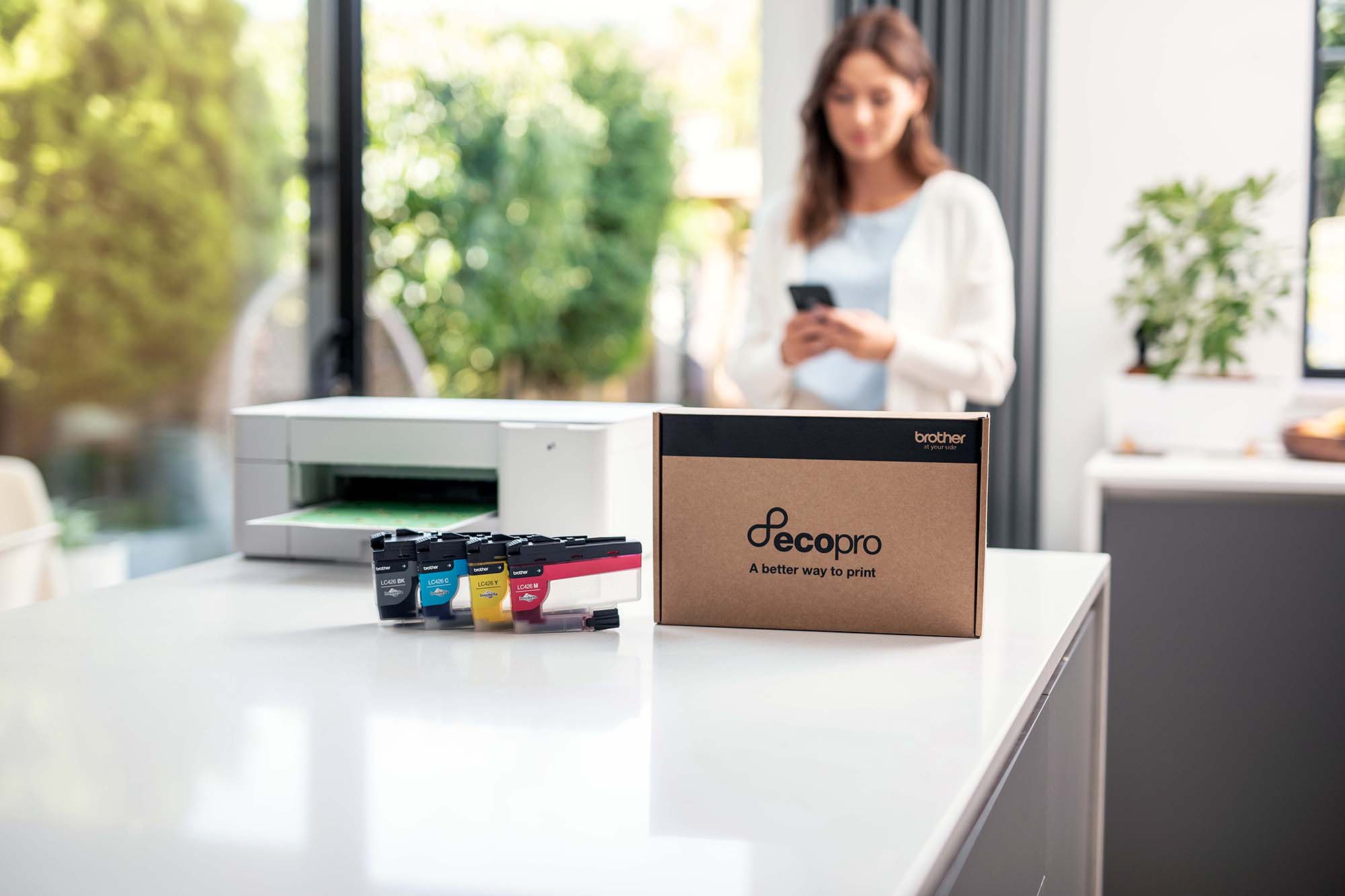 Auf einem Tisch stehen ein Drucker, Tintenpatronen in Cyan, Gelb, Magenta und Schwarz, sowie eine "ecopro"-Box. Eine Frau nutzt ein Smartphone.