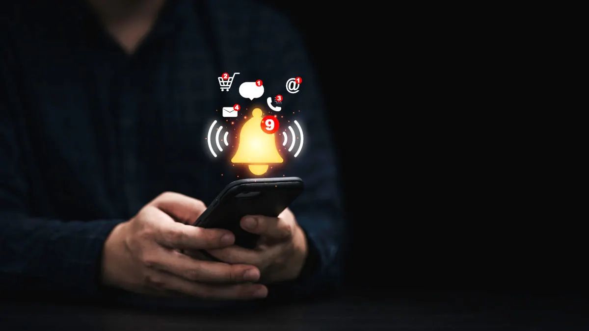 Eine Person hält ein Smartphone mit Benachrichtigungssymbolen, darunter Einkaufswagen, Sprechblase und Glocke. Der Hintergrund ist dunkel.