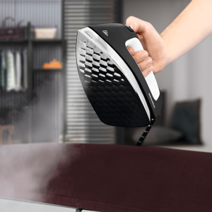 Eine Hand hält ein schwarzes Bügeleisen mit weißem Griff über einem braunen Stoff. Dampf steigt auf, ein Regal im Hintergrund.