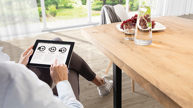 Eine Person benutzt ein Tablet mit Haus-, Zahnrad- und Schlosssymbolen neben einem Tisch mit Getränken und Trauben.