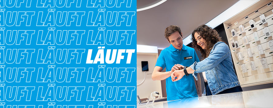 Ein Mann in blauem Hemd zeigt einer Frau eine Smartwatch in einem Geschäft. Im Hintergrund sind Handyhüllen ausgestellt. Links steht "LÄUFT".