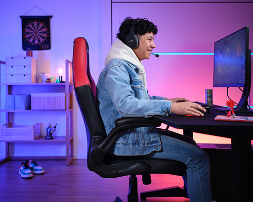 Ragazzo con cuffie gioca al computer su sedia da gaming. Scrivania nera, monitor, scaffali con oggetti e freccette sullo sfondo.