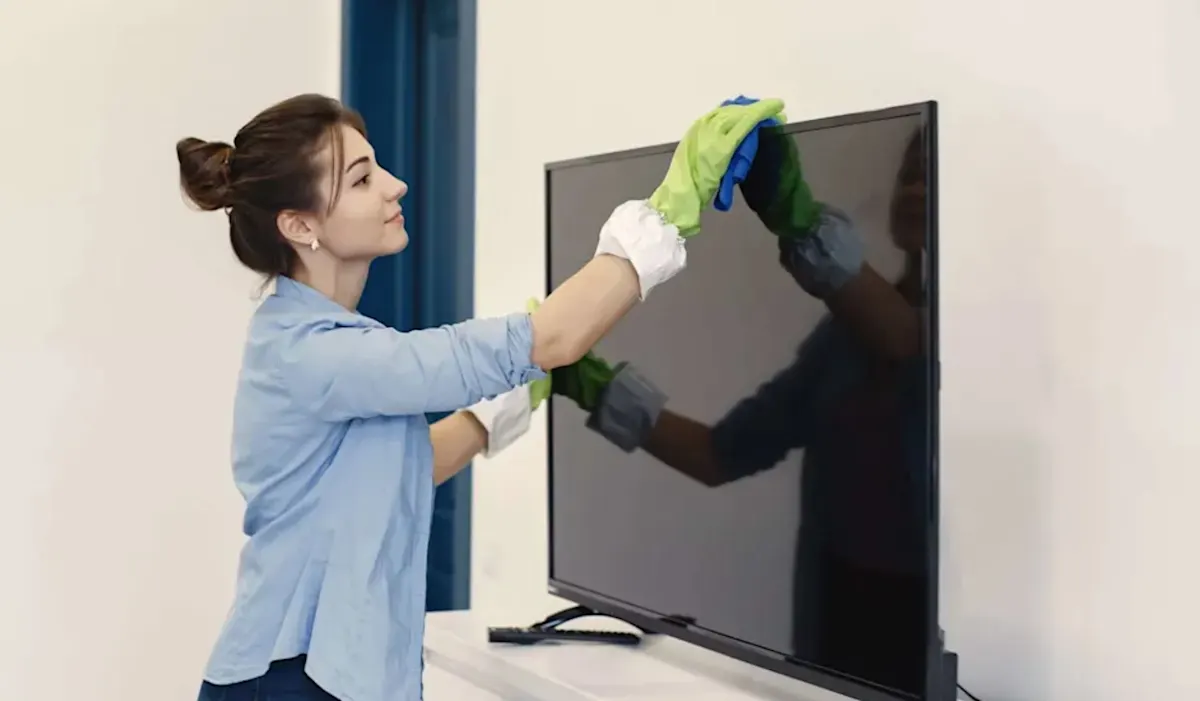 Donna che pulisce uno schermo TV nero con guanti e panno verde-azzurro in una stanza bianca.