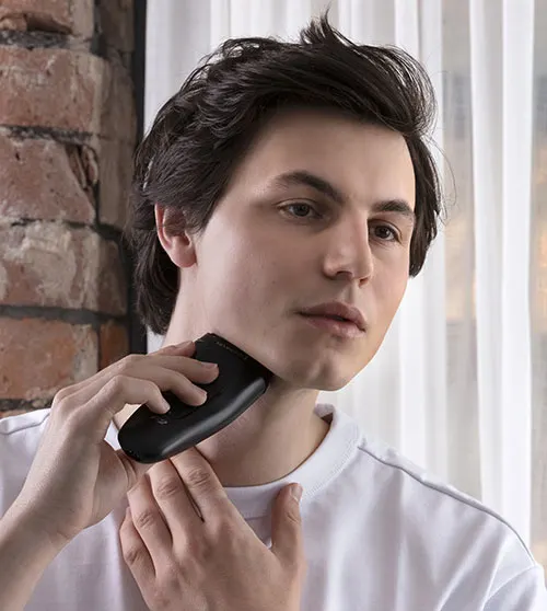 Uomo che si rade con rasoio nero su sfondo di muro di mattoni e tenda bianca. Indossa una maglietta bianca.