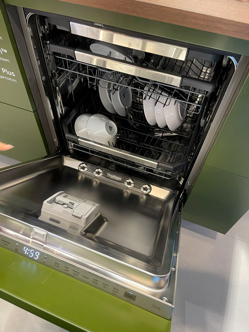 Cestello lavastoviglie aperto, ripiano superiore con tazze e piatti bianchi, ripiano inferiore vuoto con dispenser detersivo.