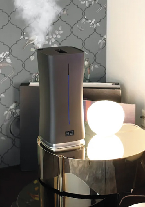 Umidificatore grigio con una striscia blu e vapore che esce dalla parte superiore, su un tavolo rotondo riflettente.