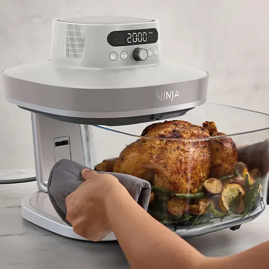 Una persona usa una presina grigia per rimuovere il grande contenitore in vetro con un pollo arrosto intero e verdure dalla friggitrice ad aria Ninja bianca.