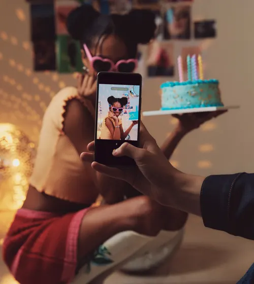 Ragazza con torta e occhiali a forma di cuore fotografata con telefono. Torta azzurra con candele. Sfondo con foto.