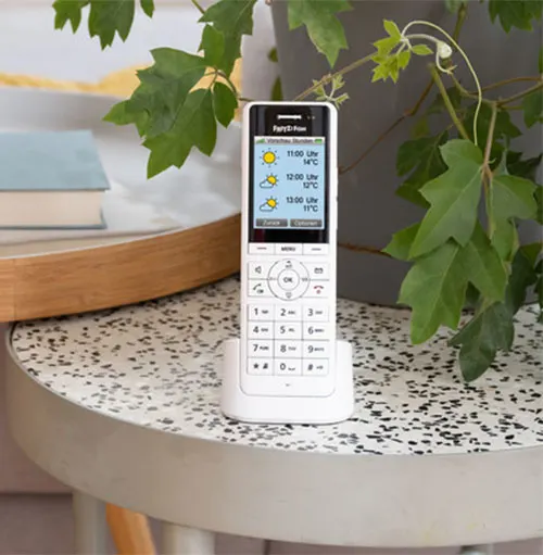 Telefono bianco su base, mostra previsioni meteo. Pianta verde sullo sfondo, tavolo rotondo con motivo a puntini.