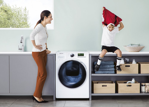 Donna accanto a lavatrice bianca. Ragazzo seduto sopra lo scaffale, tiene in mano una maglia rossa. Scatole di cartone sullo scaffale.