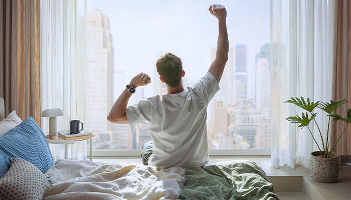 In una camera da letto c'e' un ragazzo biondo sul bordo del letto che si stira, ha un smartwatch sul polso