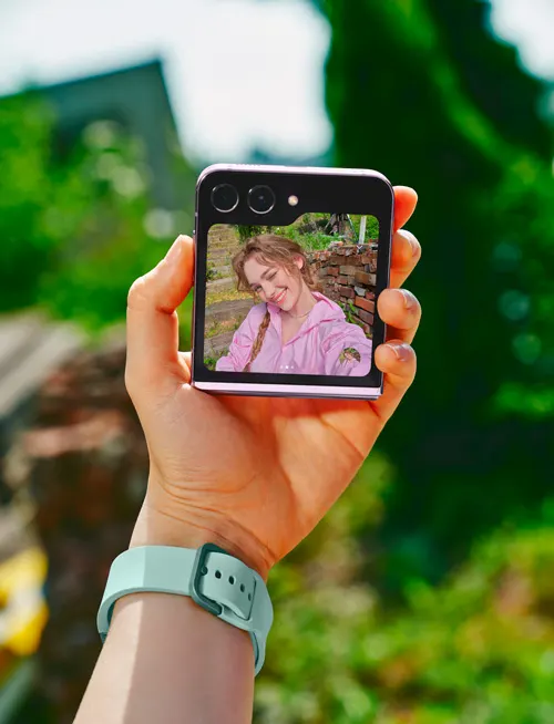 Una mano tiene un telefono che mostra una persona sorridente. Sul braccio C'� un orologio verde acqua. Lo sfondo � verde.