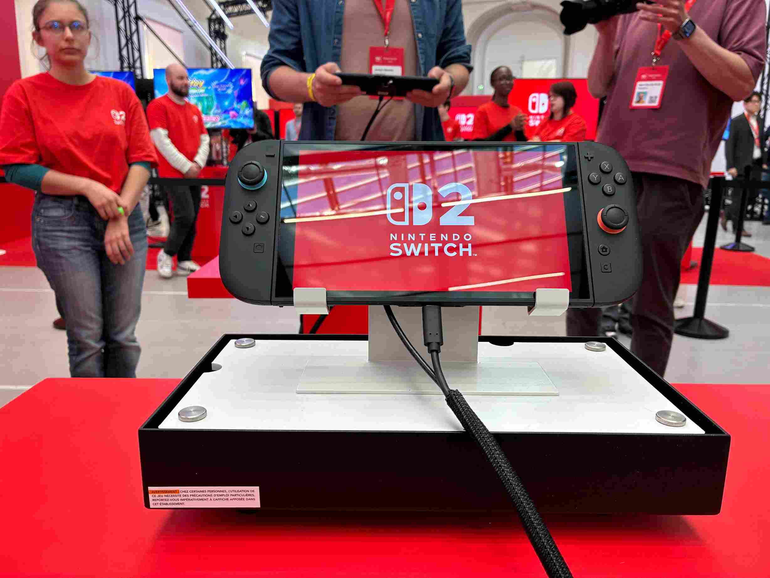 Schermo Nintendo Switch su base nera e bianca. Il display mostra il logo "Nintendo Switch". Persone sullo sfondo.