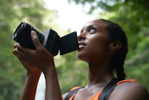 Donna che tiene in mano una fotocamera nera con uno schermo aperto, sullo sfondo alberi verdi.