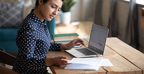 Donna seduta a una scrivania di legno con laptop, documenti e libri. Indossa una camicia blu a pois bianchi.