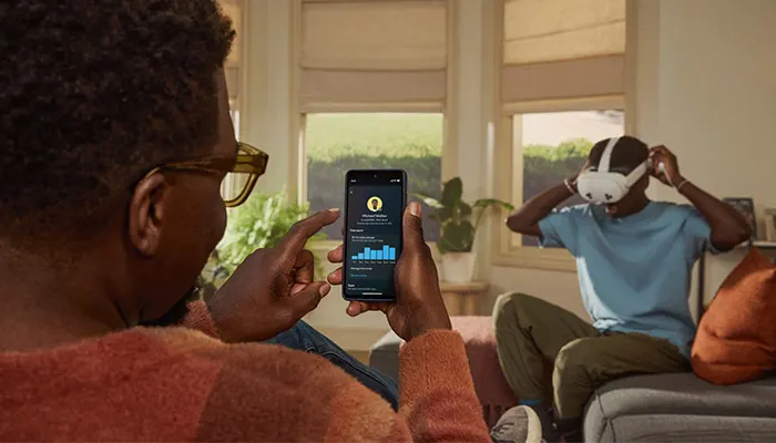 Uomo con occhiali che guarda un telefono con grafici. Un altro uomo indossa un visore VR sullo sfondo.