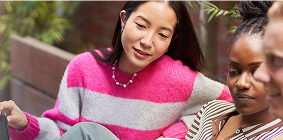 Tre persone sedute su una panchina. Una donna indossa una maglia a righe rosa e bianche e una collana di perle.