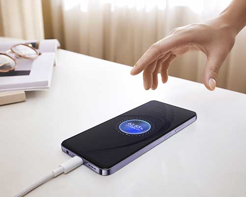 Mano sopra un telefono nero in carica su una scrivania bianca. Sullo sfondo, libri e occhiali.