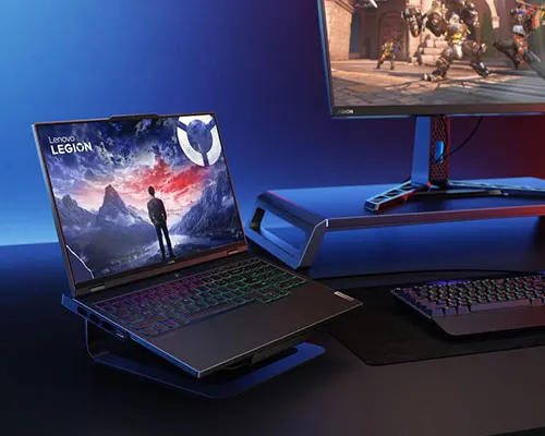 Scrivania con portatile Lenovo Legion che mostra un uomo in piedi davanti a montagne. Tastiera e monitor visibili.