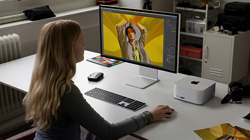 Donna seduta a una scrivania bianca con computer, tastiera, mouse e Mac Studio. Sullo schermo, un'immagine con sfondo giallo. Armadietto bianco sullo sfondo.