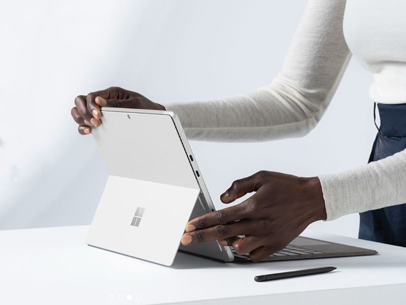 Mani regolano un tablet grigio chiaro aperto su un tavolo bianco, con una penna nera in primo piano. Sullo sfondo, una parete bianca.