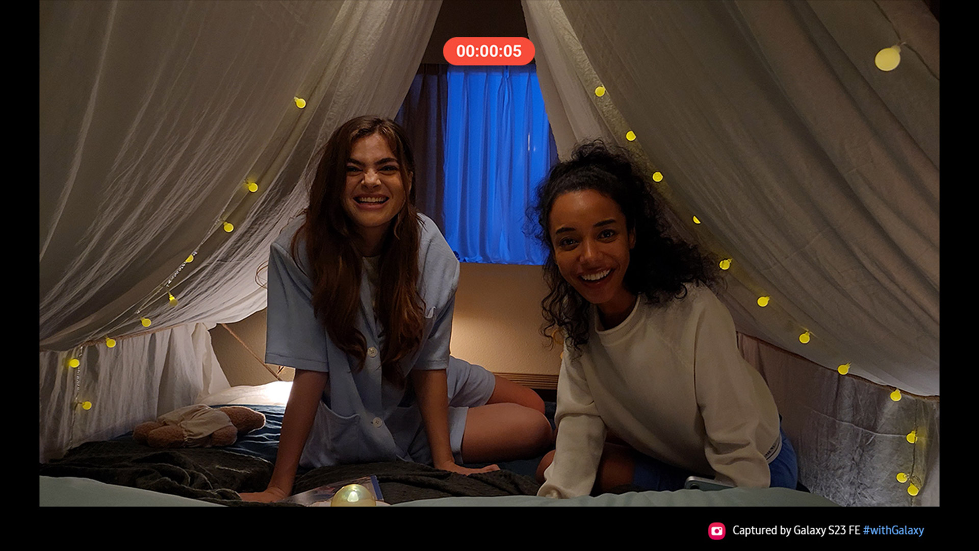 Due donne sorridono in una tenda decorata con luci. Una finestra blu � visibile sullo sfondo.