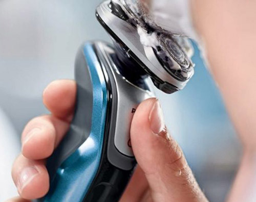 Primo piano di una mano che preme un pulsante su un rasoio elettrico blu e argento. Lo sfondo � sfocato.