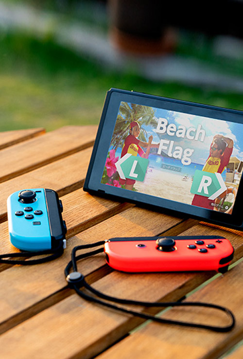 Su un tavolo di legno, una console con gioco "Beach Flag" e due controller: uno blu e uno rosso.