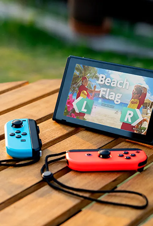 Su un tavolo di legno, una console con gioco "Beach Flag" e due controller: uno blu e uno rosso.