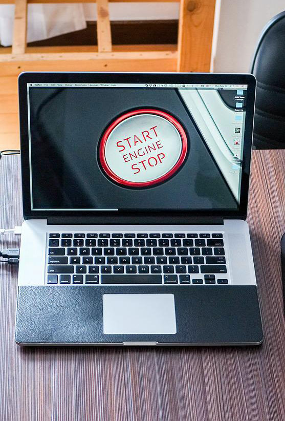 Un computer portatile aperto su una scrivania di legno mostra un'immagine di un pulsante rosso "Start Engine Stop" sullo schermo.