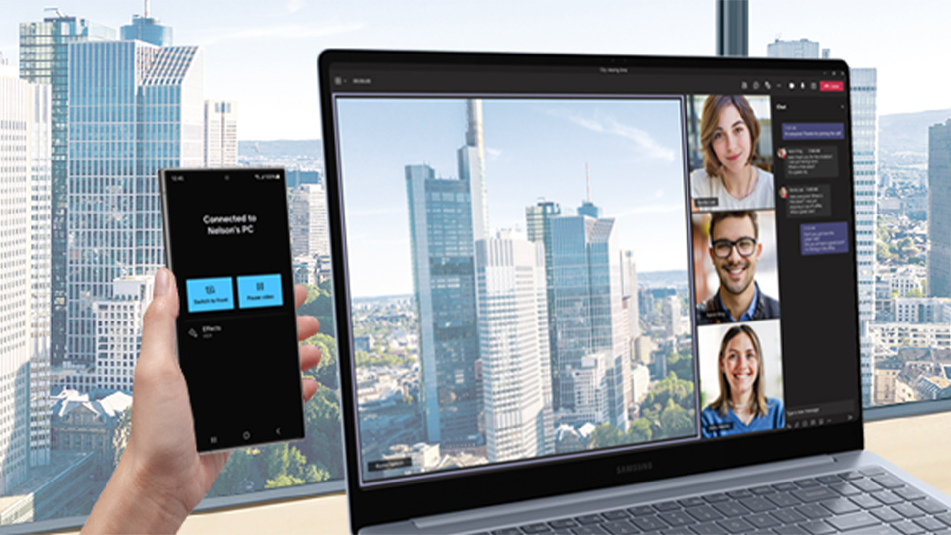 Schermo di laptop mostra videochiamata con tre persone; mano tiene telefono connesso a PC. Grattacieli sullo sfondo.