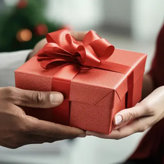 Mani che offrono un regalo rosso con nastro e fiocco, sfondo sfocato con albero decorato.