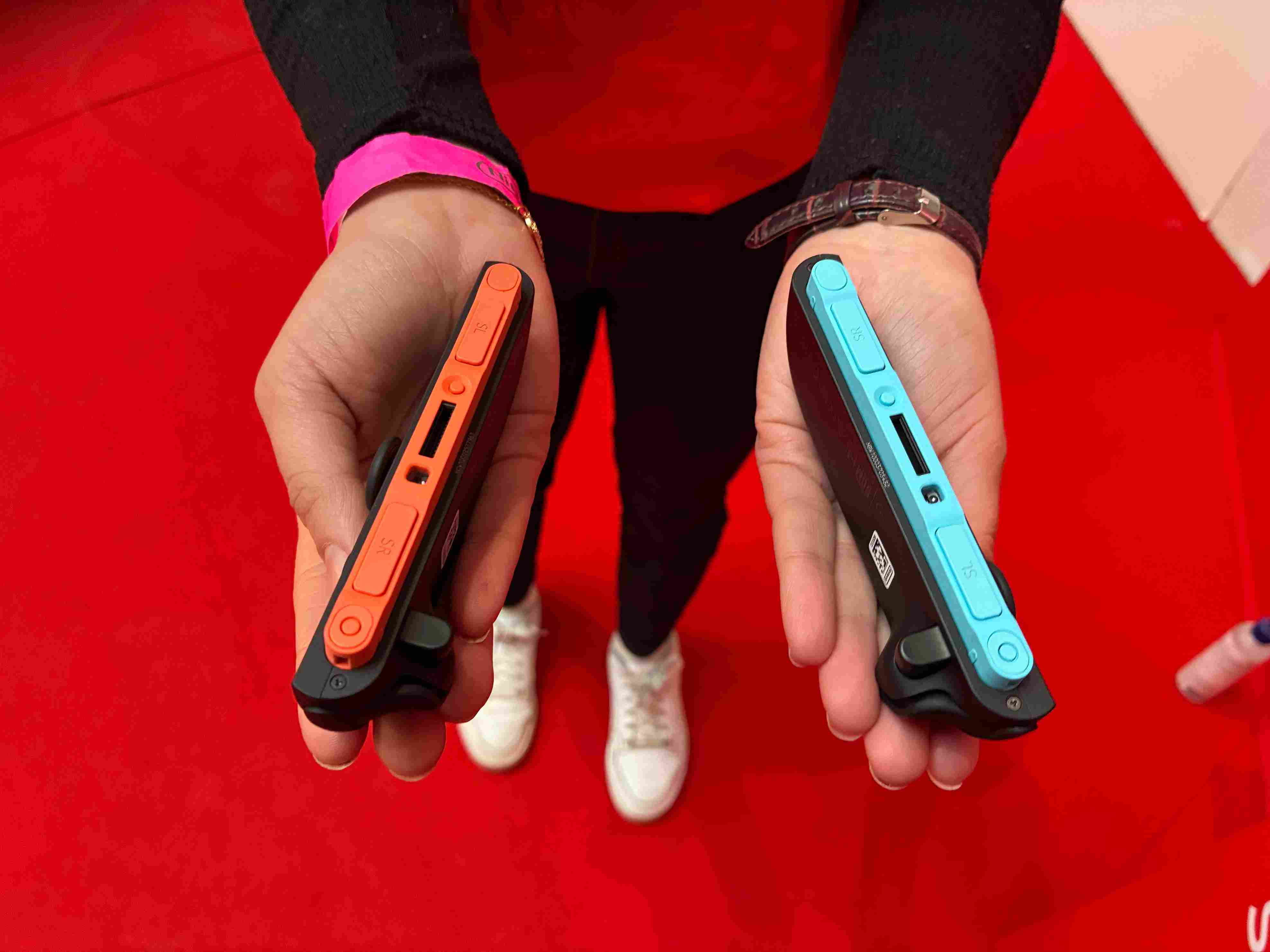 Due dispositivi neri con bordi arancioni e azzurri tenuti su sfondo rosso. Una persona indossa scarpe bianche.