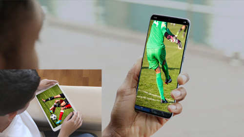 Un uomo guarda una partita di calcio su un telefono e un tablet. Il gioco � su erba verde.
