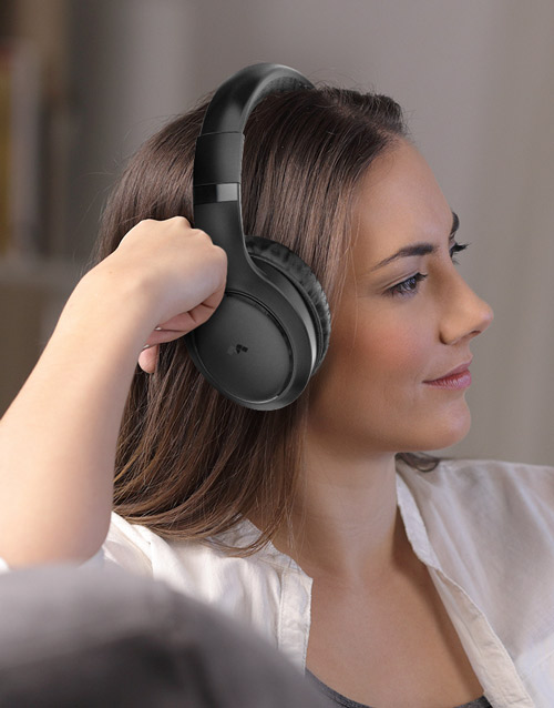 Donna con cuffie nere over-ear, indossa camicia bianca in un interno con sfondo neutro. Mano alzata verso la cuffia.