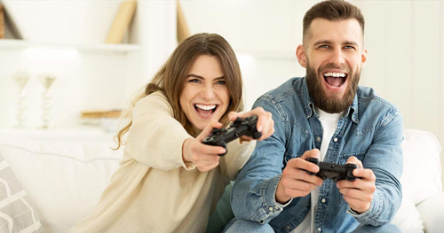 Una coppia sorride mentre gioca ai videogiochi con i controller. Sono seduti su un divano bianco in una stanza luminosa.