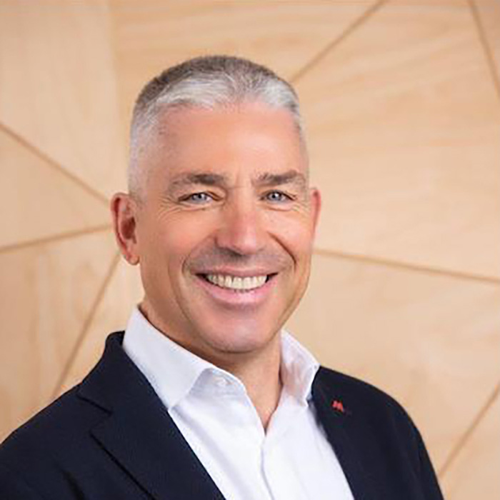 Uomo sorridente in giacca blu e camicia bianca, su sfondo di pannelli di legno chiaro.