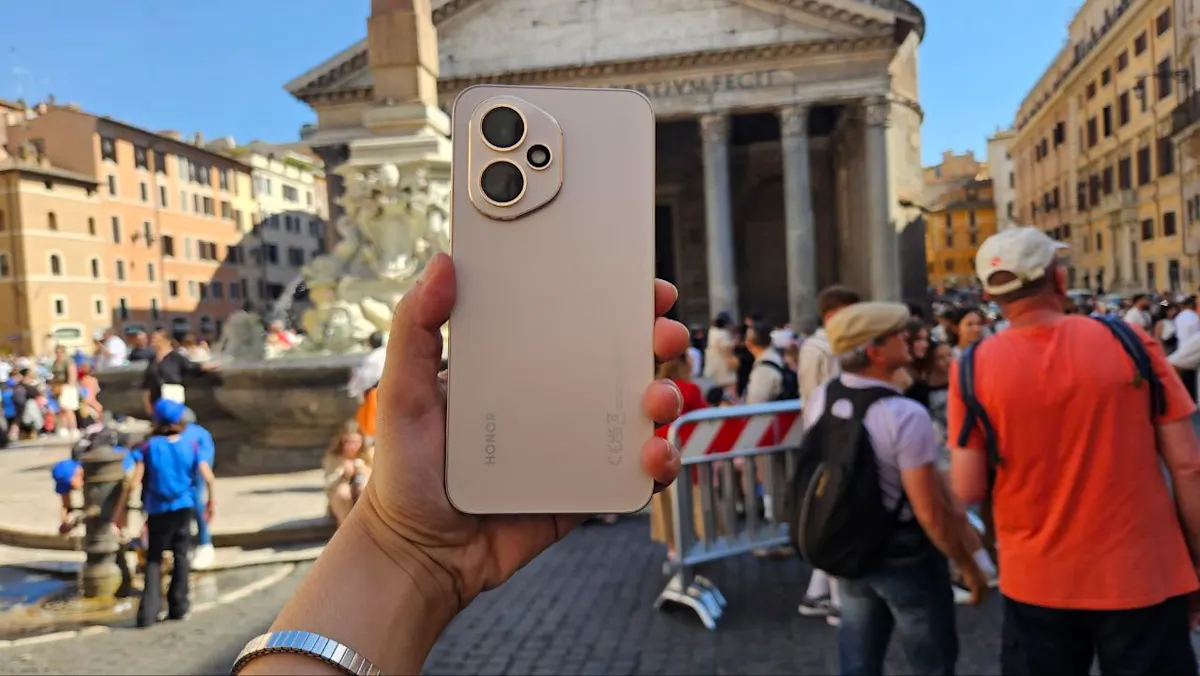 Una mano tiene un telefono color crema. Sullo sfondo, un edificio antico e persone in una piazza affollata.